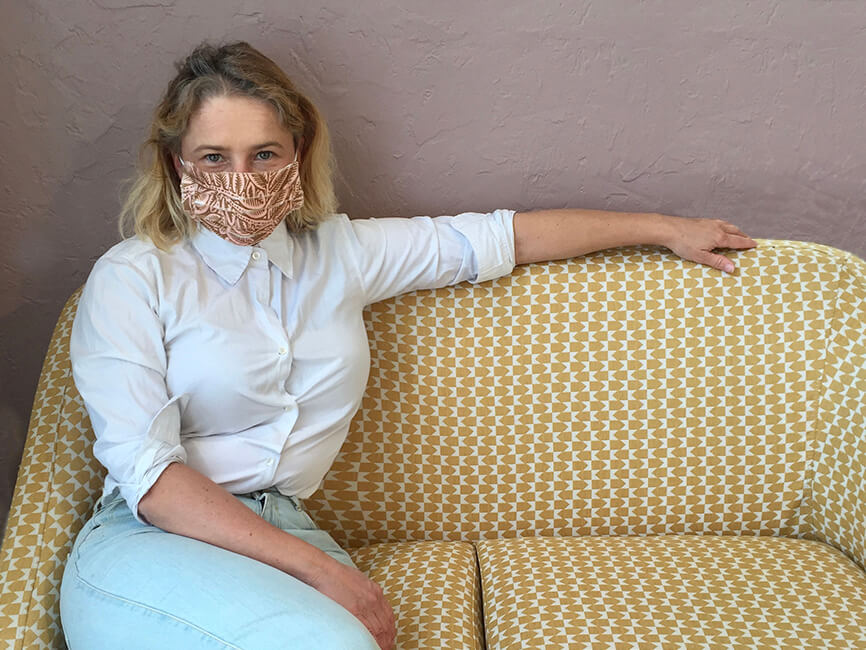 lady sitting on a sofa, wearing white shirt and pink disposable mask on her face
