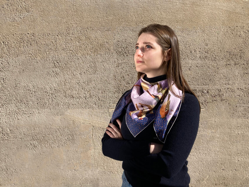 jeune femme portant un pull marine et un foulard en soie mauve devant un mur