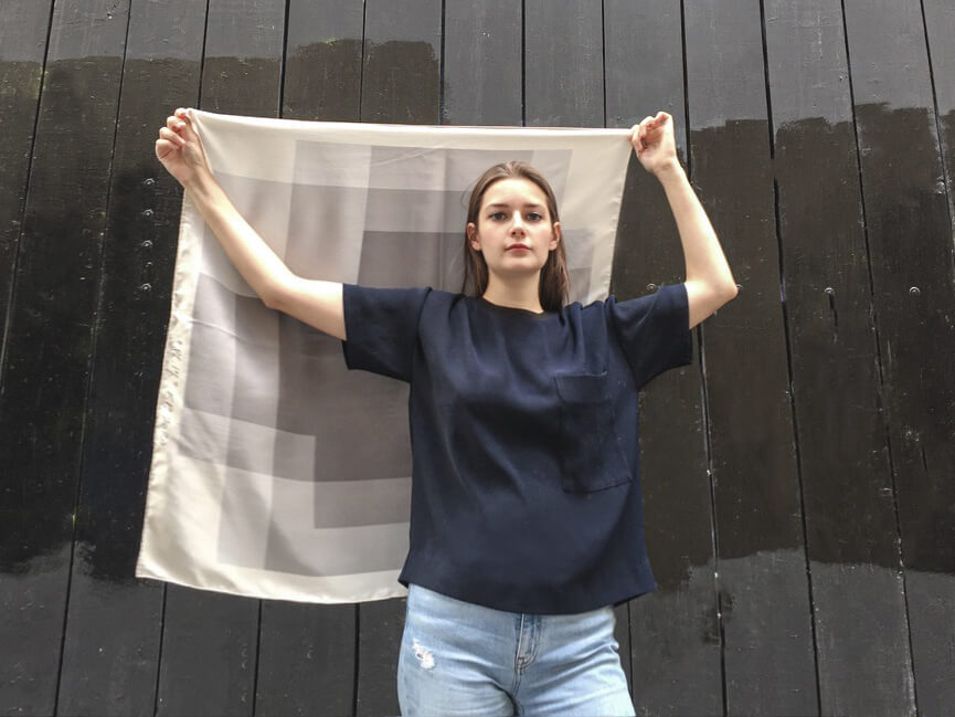 young lady wearing jean and holding in her back a grey scarf ith black wall in the background