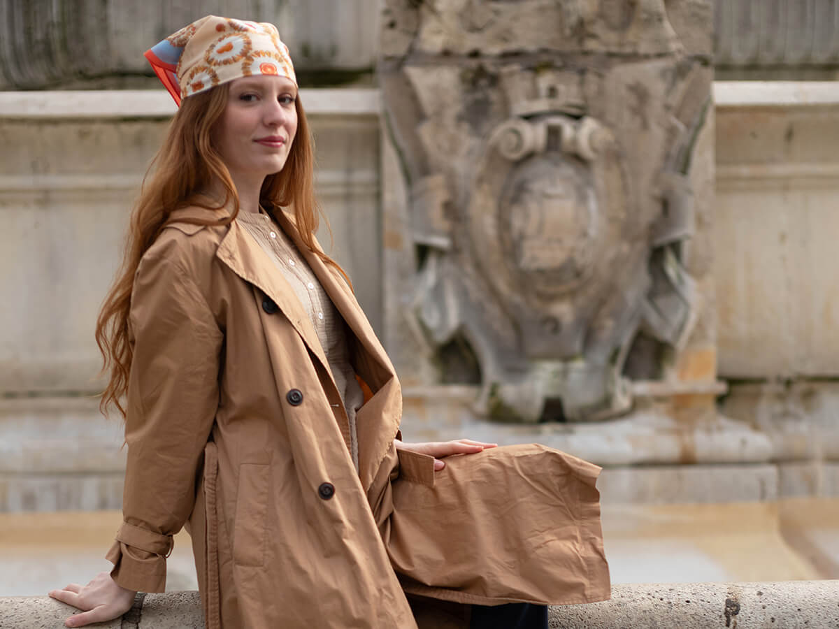 jeune femme rousse avec un foulard en coton et soie sur la tête et un manteau beige assise devant une fontaine