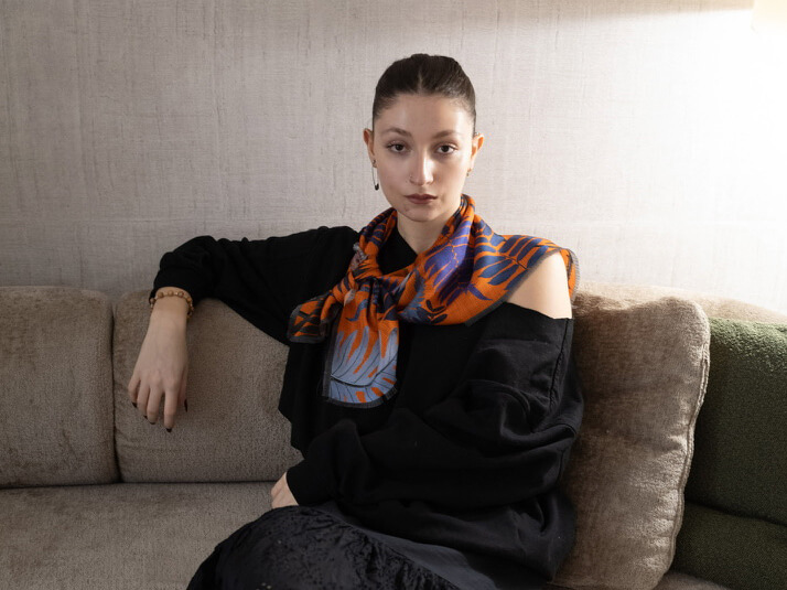 white woman wearing a black pull over and an orange silk scarf with leaves motif around her neck sitting on a couch