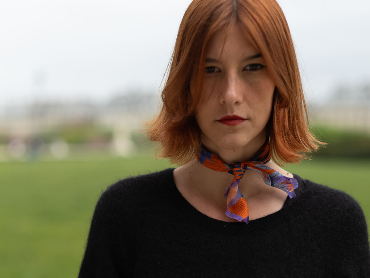 white woman wearing a black pull over and an orange silk scarf with leaves motif around her neck