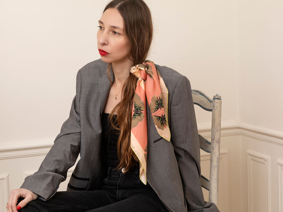 white woman wearing a pink silk scarf with butterfly motif with a grey jacket and a black dress sitting on a chair