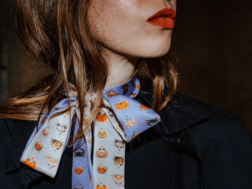 white woman wearing a purple long skinny silk scarf with crab motif tied around her neck and a black jacket